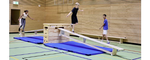 Kinder in Sporthalle bei einem Parkour aus Turnbänken, Sprungkästen und Matten